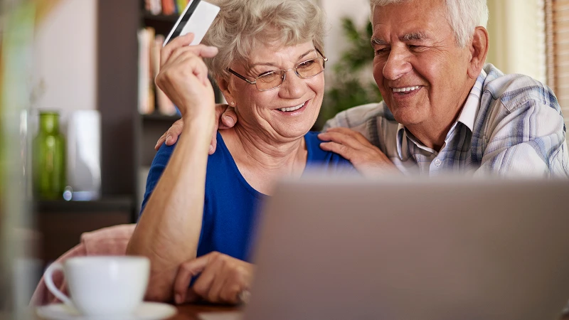 Smiling retired couple looking at a laptop and holding a credit card, representing the process of paying off high-interest debt using home equity.